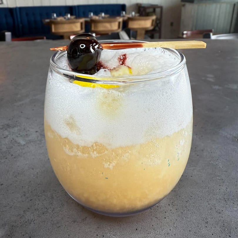 A Comida cocktail with a foamy top, garnished with a dark cherry and a lemon slice.
