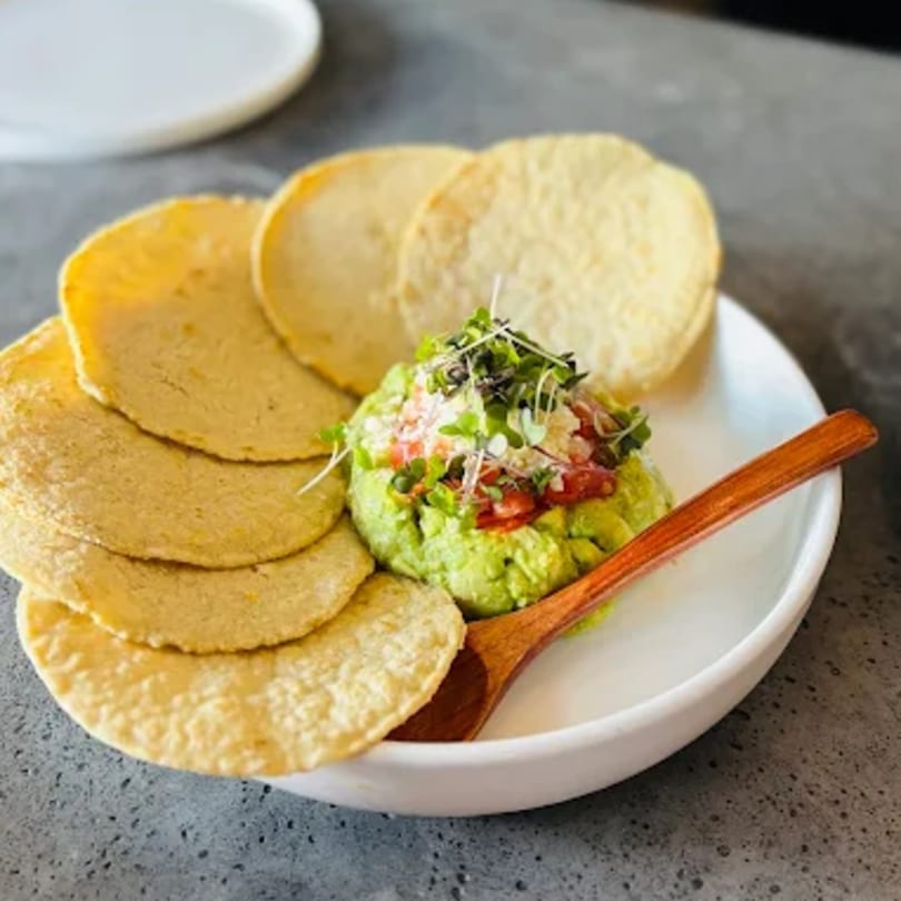 Comida's guacamole with microgreens, tomatoes, and cheese, served with corn tortillas and a wooden spoon.